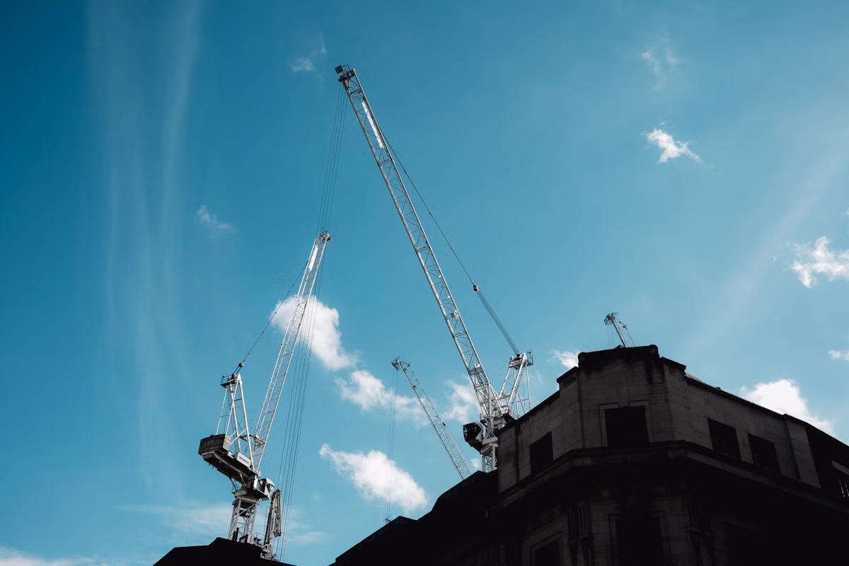 Tower cranes rise against a blue sky behind the roofline of a stone-faced city building.