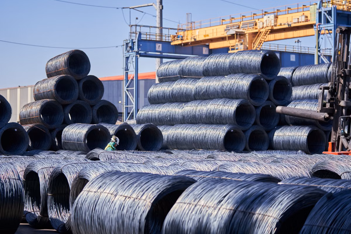 Stacked coils of steel wire rod in an outdoor storage yard beneath an overhead gantry crane, with a worker in a hard hat and high-visibility clothing among the stacks.