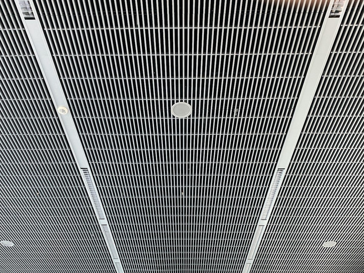 Looking up at a suspended open-grid metal ceiling with linear light strips, a smoke detector and a round speaker fitted between the panels.
